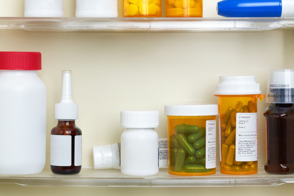 A medicine cabinet shelf containing various prescription bottles, pill containers, a nasal spray, and a brown bottle with a dropper—just like what a veterinarian or vet might use—all with blank or partially visible labels.